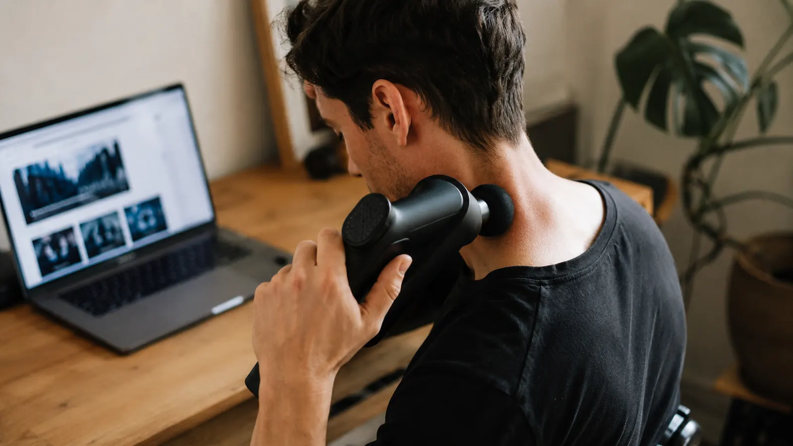 Person using a massage gun on the side of their neck and upper trap area while sitting at a desk, laptop visible in the background