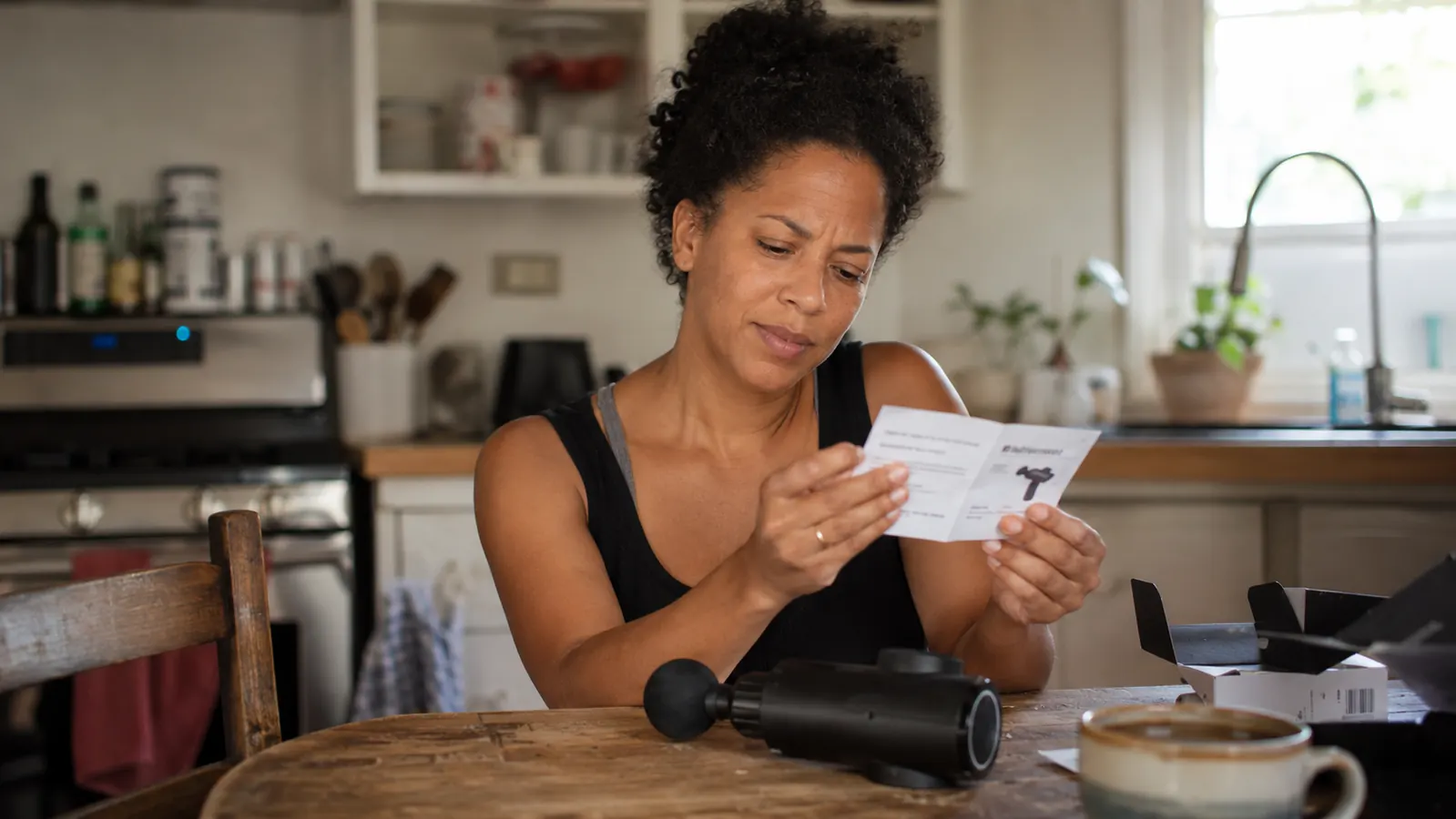 Woman in her late 30s sitting at a kitchen table with a RENPHO massage gun in front of her, arms crossed, reading the instruction manual with a skeptical expression